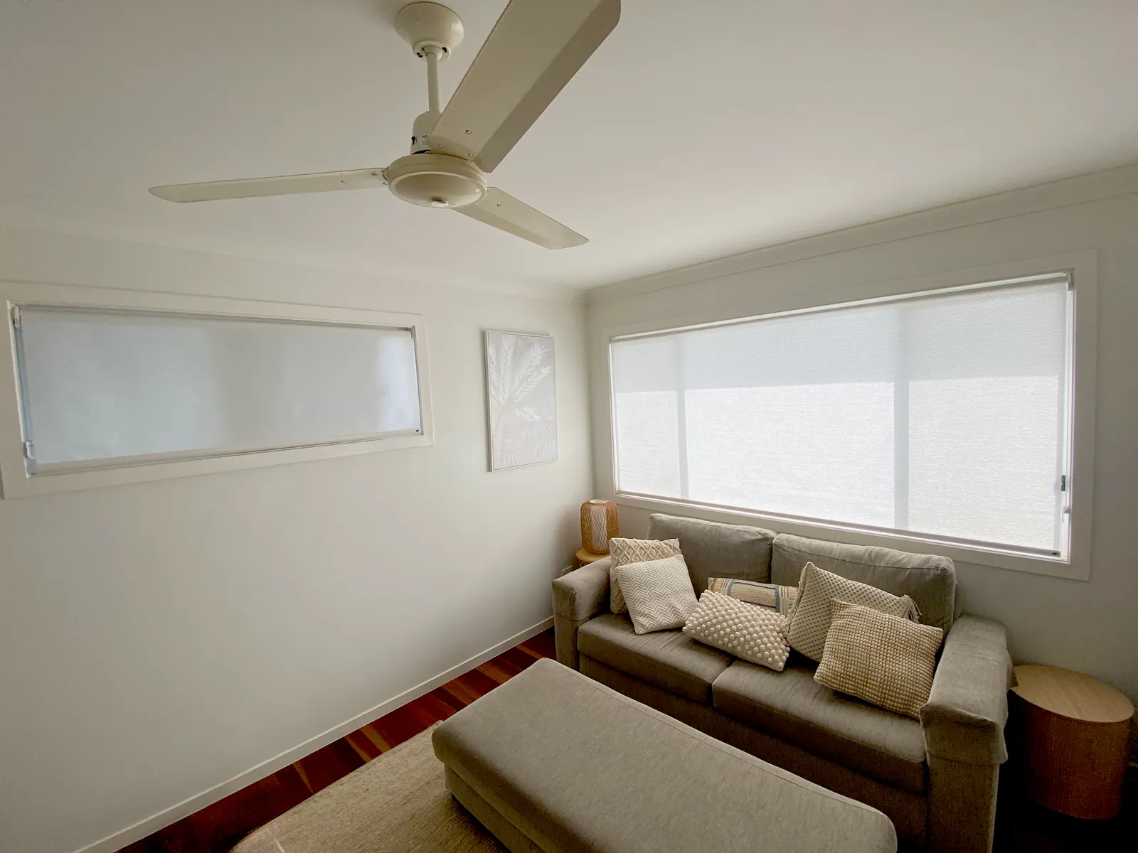 A lounge room with two white roller blinds down over long windows, a grey sofa and a ceiling fan.